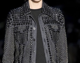 May include: A dark gray denim jacket heavily embellished with black studs and buttons. The jacket has a classic collar, two chest pockets, and a button-down front. The jacket is worn over a black dress.