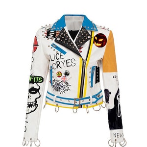Könnte beinhalten: Weiße Lederjacke mit blauem Kragen und gelben Akzenten. Die Jacke ist mit verschiedenen Patches, Pins und grafischen Designs verziert, darunter ein Totenkopf und Text. Silberne Metallnieten und Ringe verstärken den Punk-Rock-Look. Der Text "Lice Oryes" ist sichtbar.
