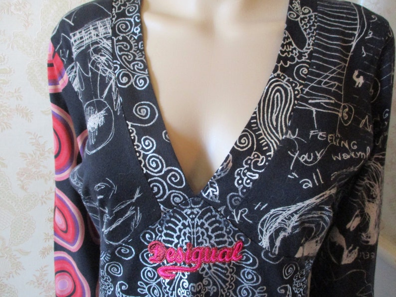 May include: Black long-sleeve shirt with a silver and white abstract print. The shirt has a V-neck and a pink "Desigual" logo on the front.