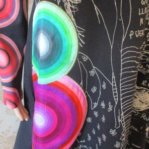 May include: Black tunic dress with a colorful, concentric circle design on the side. The circles are in shades of pink, purple, green, and blue. The dress has a white, hand-drawn design on the front.