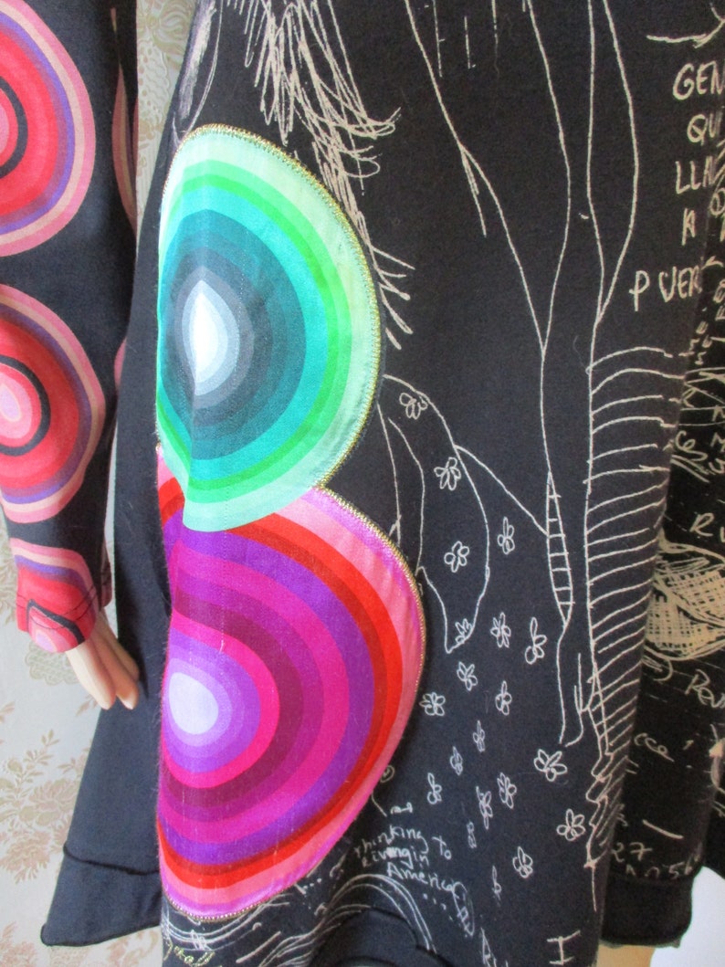 May include: Black tunic dress with a colorful, concentric circle design on the side. The circles are in shades of pink, purple, green, and blue. The dress has a white, hand-drawn design on the front.
