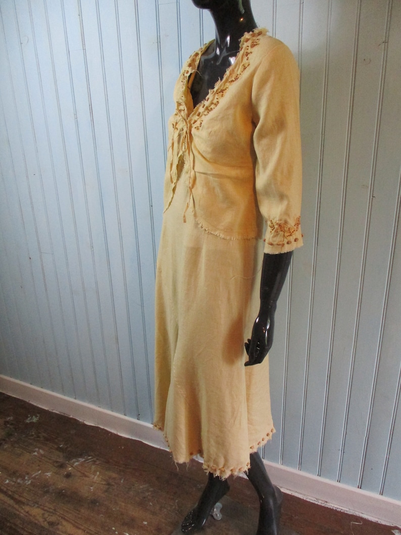 May include: A light yellow dress with a long skirt and a short-sleeved top. The dress has a V-neckline and is decorated with brown embroidery around the neckline and sleeves.