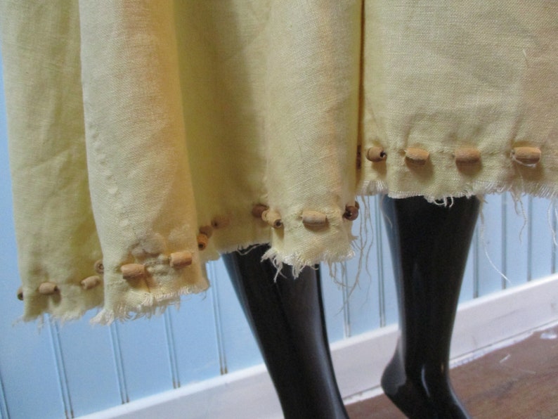 May include: A pale yellow linen skirt with a raw edge hem. The skirt is decorated with a row of wooden beads.