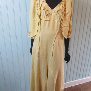 May include: A long, pale yellow linen dress with a matching jacket. The dress has a V-neckline and a flowing skirt. The jacket has a scalloped hem and is decorated with brown beading.