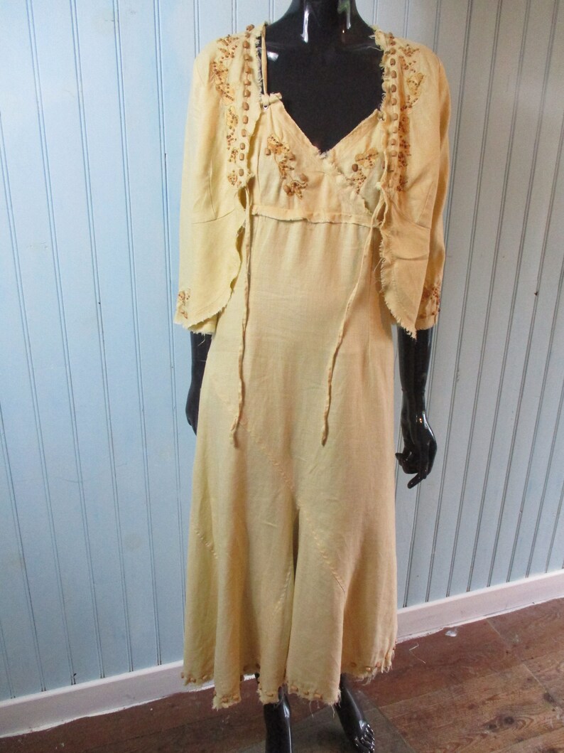 May include: A long, pale yellow linen dress with a matching jacket. The dress has a V-neckline and a flowing skirt. The jacket has a scalloped hem and is decorated with brown beading.
