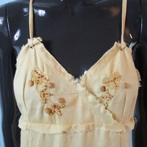 May include: A light yellow sleeveless dress with a V-neckline and a frayed hem. The dress is decorated with embroidered floral designs and brown wooden beads.