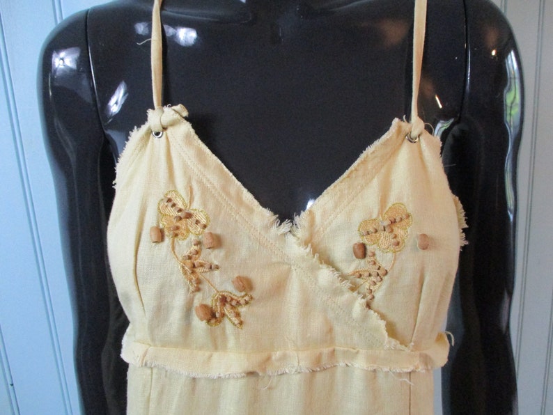 May include: A light yellow sleeveless dress with a V-neckline and a frayed hem. The dress is decorated with embroidered floral designs and brown wooden beads.