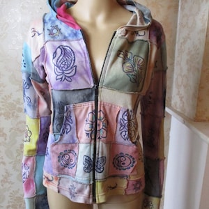 Könnte beinhalten: Eine Patchwork-Kapuzenjacke mit Reißverschluss. Die Jacke besteht aus verschiedenen Stoffen in unterschiedlichen Farben mit verschiedenen Designs, darunter Blumen, Paisley-Muster und Yin-Yang-Symbole.