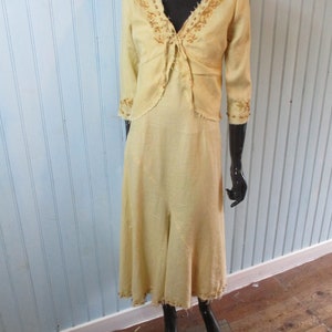 May include: A yellow linen dress with a matching embroidered jacket. The dress has a V-neckline and a flowing skirt. The jacket has a tie closure and three-quarter sleeves.