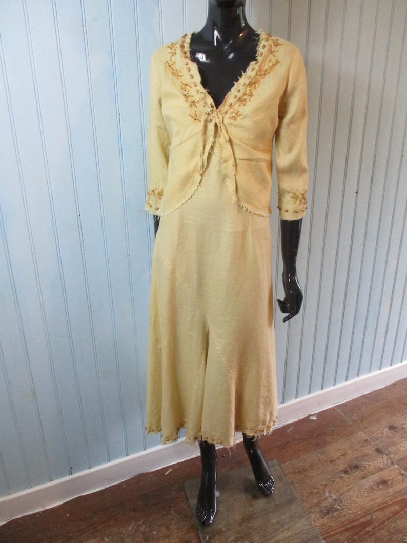 May include: A yellow linen dress with a matching embroidered jacket. The dress has a V-neckline and a flowing skirt. The jacket has a tie closure and three-quarter sleeves.