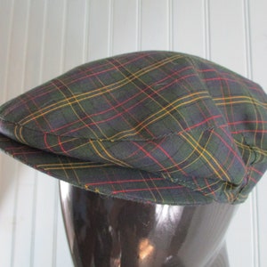 May include: A green, navy, red, and yellow plaid flat cap. The hat is made of a woven fabric and has a rounded crown and a short, stiff peak. The cap is displayed on a mannequin head.