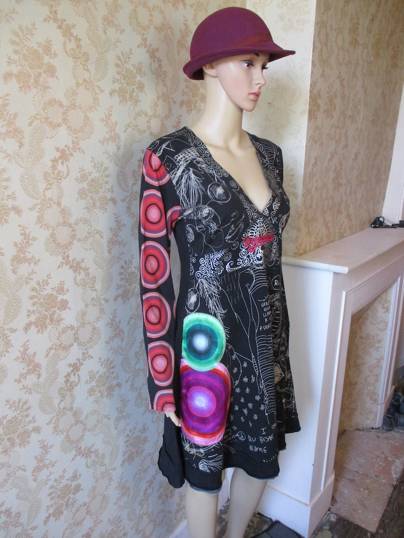 May include: A black and white patterned dress with a red, pink, and green circle design on the sleeve. The dress has a V-neckline and a burgundy hat.