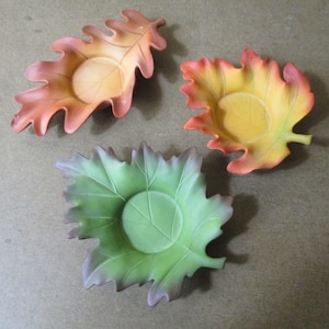 May include: Three ceramic leaf-shaped candle holders. The holders are in the shape of oak leaves. The leaves are in shades of orange, yellow, and green.