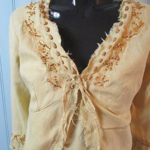 May include: A light yellow linen jacket with a tie front and brown bead embroidery. The jacket has a frayed neckline and cuffs.