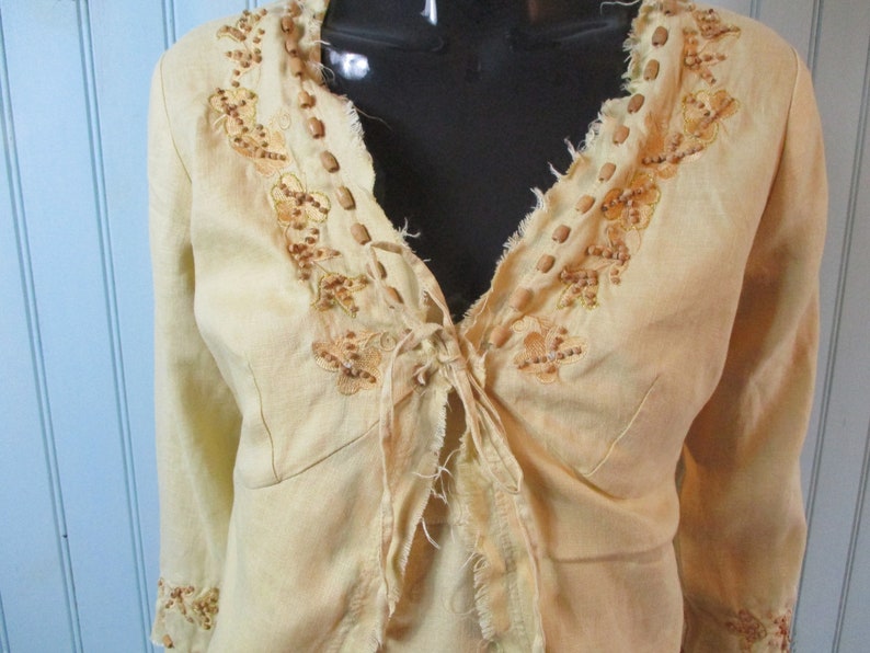 May include: A light yellow linen jacket with a tie front and brown bead embroidery. The jacket has a frayed neckline and cuffs.