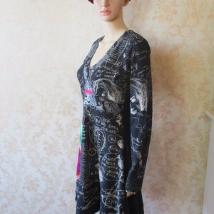 May include: Black dress with long sleeves and a V-neck. The dress has a white print of scribbles, words, and drawings. The model is wearing a burgundy fedora.