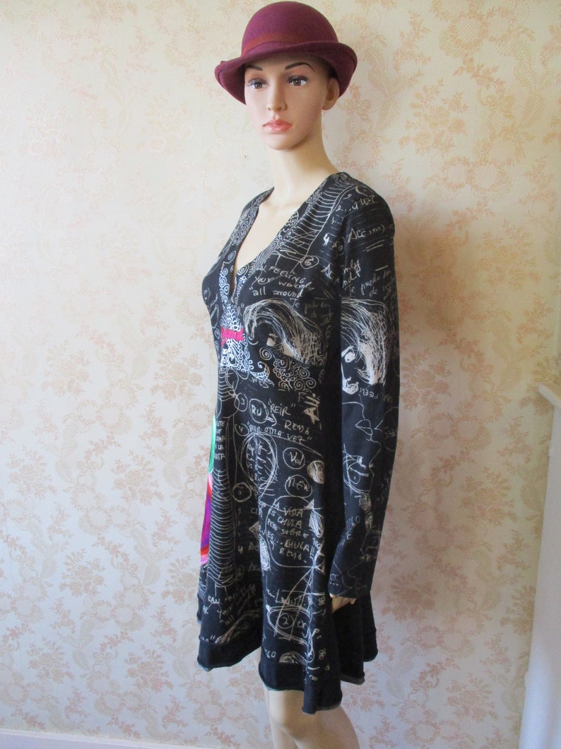 May include: Black dress with long sleeves and a V-neck. The dress has a white print of scribbles, words, and drawings. The model is wearing a burgundy fedora.