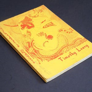 Könnte beinhalten: Ein Vintage-Buch mit gelbem Einband mit roten Illustrationen und Text. Der Titel "Psychedelic Prayers" ist vertikal entlang des Buchrückens gedruckt, mit "Timothy Leary" in Rot auf dem vorderen Einband. Das Buch ist vor einem dunklen Hintergrund platziert.
