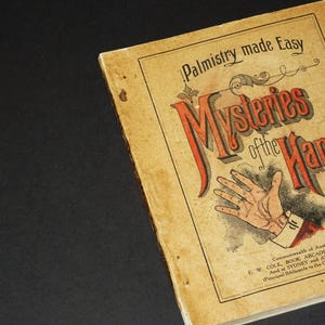 Op de afbeelding: Een vintage boek getiteld "Palmistry made Easy Mysteries of the Hand" met een illustratie van een hand. De boekomslag is verkleurd door de tijd en heeft een rode en zwarte titel.