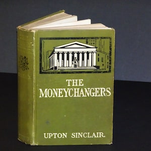 Pode incluir: Um livro de capa dura verde com o título "THE MONEYCHANGERS" de UPTON SINCLAIR. A capa apresenta uma ilustração em preto e branco de um edifício com colunas.