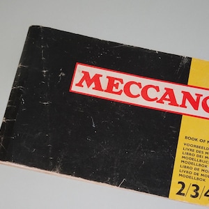 May include: A black and yellow book with the title "Meccano Book of Models" and the numbers "2/3/4" printed on the cover. The book has multiple languages listed on the cover, including English, French, German, Italian, Spanish, and Dutch.