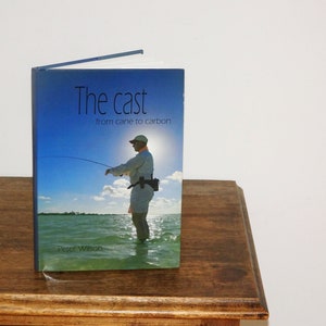 Könnte beinhalten: Ein Buch mit dem Titel "The cast from cane to carbon" von Peter Wilson. Das Cover zeigt ein Foto einer Person, die in einem türkisfarbenen Gewässer mit blauem Himmel und weißen Wolken im Hintergrund Fliegenfischen betreibt.