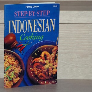 Op de afbeelding: Een kookboek getiteld "Family Circle Step-By-Step Indonesian Cooking" met een blauwe omslag. De omslag toont afbeeldingen van Indonesische gerechten, waaronder garnalen en rijst. De titel van het boek staat in grote, vette letters.
