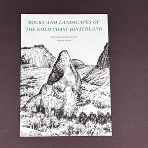 May include: A softcover book titled Rocks and Landscapes of the Gold Coast Hinterland, Expanded Second Edition by Warwick Willmott. The white cover features a black and white ink illustration of a prominent mountain peak and surrounding forest.