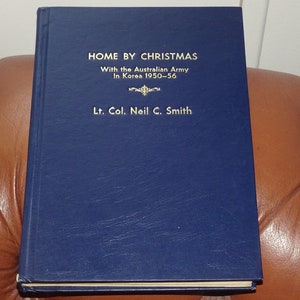 May include: A navy blue hardback book with gold lettering. The title is "Home by Christmas" with the subtitle "With the Australian Army in Korea 1950-56". The author is "Lt. Col. Neil C. Smith".
