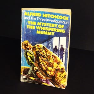 May include: Vintage paperback book titled "Alfred Hitchcock and The Three Investigators in The Mystery of the Whispering Mummy." The cover features a mummy, figures, and text on a blue background.
