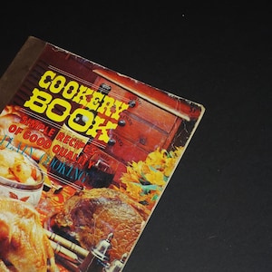 May include: A vintage cookbook with a red, orange and yellow cover. The cover features a photo of a roasted turkey, a ham and a bowl of potatoes. The title of the book is "Cookery Book: Simple Recipes of Good Quality".