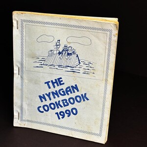 May include: A vintage cookbook titled "The Nyngan Cookbook 1990." The cover features a blue illustration of people on a raft, with a decorative border. The book has a worn appearance, suggesting it is a used item.