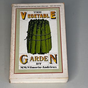 May include: A hardback book titled The Vegetable Garden by MM. Vilmorin-Andrieux. The white cover features a central illustration of a bundle of green asparagus tied with string, framed by a green border, with the text "Nearly one hundred years later this book remains without equal..." EAST/WEST JOURNAL at the top.
