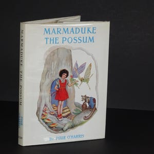 May include: A book cover with a white background and the title "Marmaduke the Possum" in black text. The cover features an illustration of a girl in a red dress standing in front of a tree with a possum and fairies.