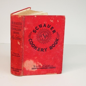 以下が含まれることがあります： 黒い文字で書かれた、ヴィンテージの赤い料理本「Schauer Australian Cookery Book」。本は開かれており、古くなったページが見えます。表紙には円形のデザインで「Schauer Cookery Book」と「Tenth Edition」と書かれています。