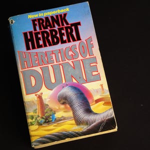 May include: A paperback book titled "Heretics of Dune" by Frank Herbert. The cover features a futuristic landscape with a large, segmented tube-like structure, a pink planet, and the text "New in paperback".