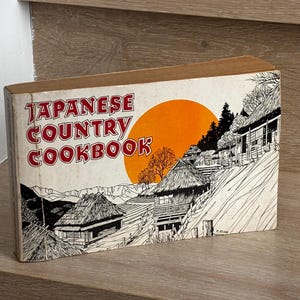 May include: A rectangular cookbook titled Japanese Country Cookbook with a white cover featuring a black ink illustration of rural houses and a large orange sun. The text is printed in red with a black outline on the left side of the cover.