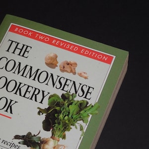 May include: The cover of a green cookbook titled "The Commonsense Cookery Book" with the subtitle "Book Two Revised Edition". The cover features a bunch of green leafy vegetables and some mushrooms.