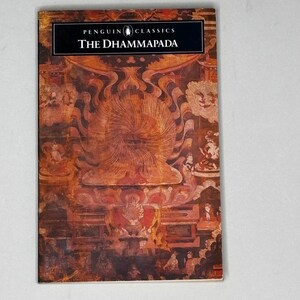 May include: A paperback book titled The Dhammapada from the Penguin Classics series. The cover features a detailed, warm-toned illustration of a central radiating sun-like motif surrounded by smaller figures in a traditional artistic style.