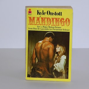 Puede incluir: Una cubierta de libro de bolsillo amarilla con el título "MANDINGO" en letras rojas grandes y en negrita. El nombre del autor, "Kyle Onstott", está impreso en letras negras sobre el título. La cubierta presenta una foto en blanco y negro de un hombre y una mujer. El texto "Now a Major Motion Picture from Dino De Laurentiis for Paramount Release" está impreso en letras negras debajo del título.