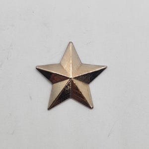 May include: A bronze-coloured star-shaped decorative element. The star has five points and a textured surface, reflecting light. It is likely a decorative pin or embellishment, suitable for various craft projects or as an accessory.