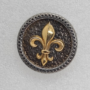 Könnte beinhalten: Ein runder, silberfarbener Knopf mit einer strukturierten Oberfläche und einem goldfarbenen Fleur-de-lis-Motiv.