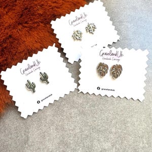 May include: Three pairs of silver-toned stud earrings displayed on white scalloped cards labelled Graceland Uk Handmade Earrings. One pair features cactus shapes, one pair features maple leaf shapes, and one pair features monstera leaf shapes.