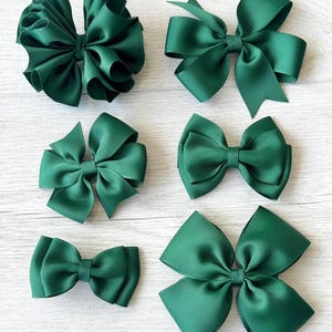 May include: Six dark forest green hair bows made of ribbed grosgrain ribbon. The collection includes various styles, ranging from a multi-layered ruffled bow to classic double-loop designs and a simple, smaller bow, all featuring a consistent texture.