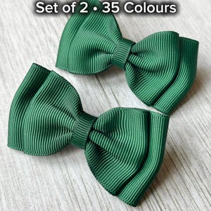 May include: Two dark green hair bows with a ribbed texture are shown on a light wood-effect surface. The bows are tied in a classic bow shape. Text at the top reads "Set of 2 • 35 Colours".