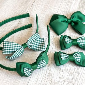 May include: A selection of green hair accessories, including two headbands and three bow clips. The headbands feature green gingham bows, while the clips have solid green and gingham bows. The accessories are displayed on a light-coloured wooden surface.