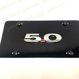 May include: Black metal license plate frame with a chrome 5.0 emblem and a red accent.