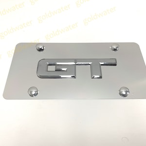 May include: A silver metal license plate frame with the letters "GT" in raised chrome lettering.