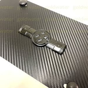 May include: Black carbon fiber license plate frame with a black metal emblem featuring the Toyota logo. The frame has four black plastic mounting points.