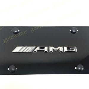 May include: Black license plate with the chrome letters "AMG" in the center. The plate has four black mounting studs. The background is white.